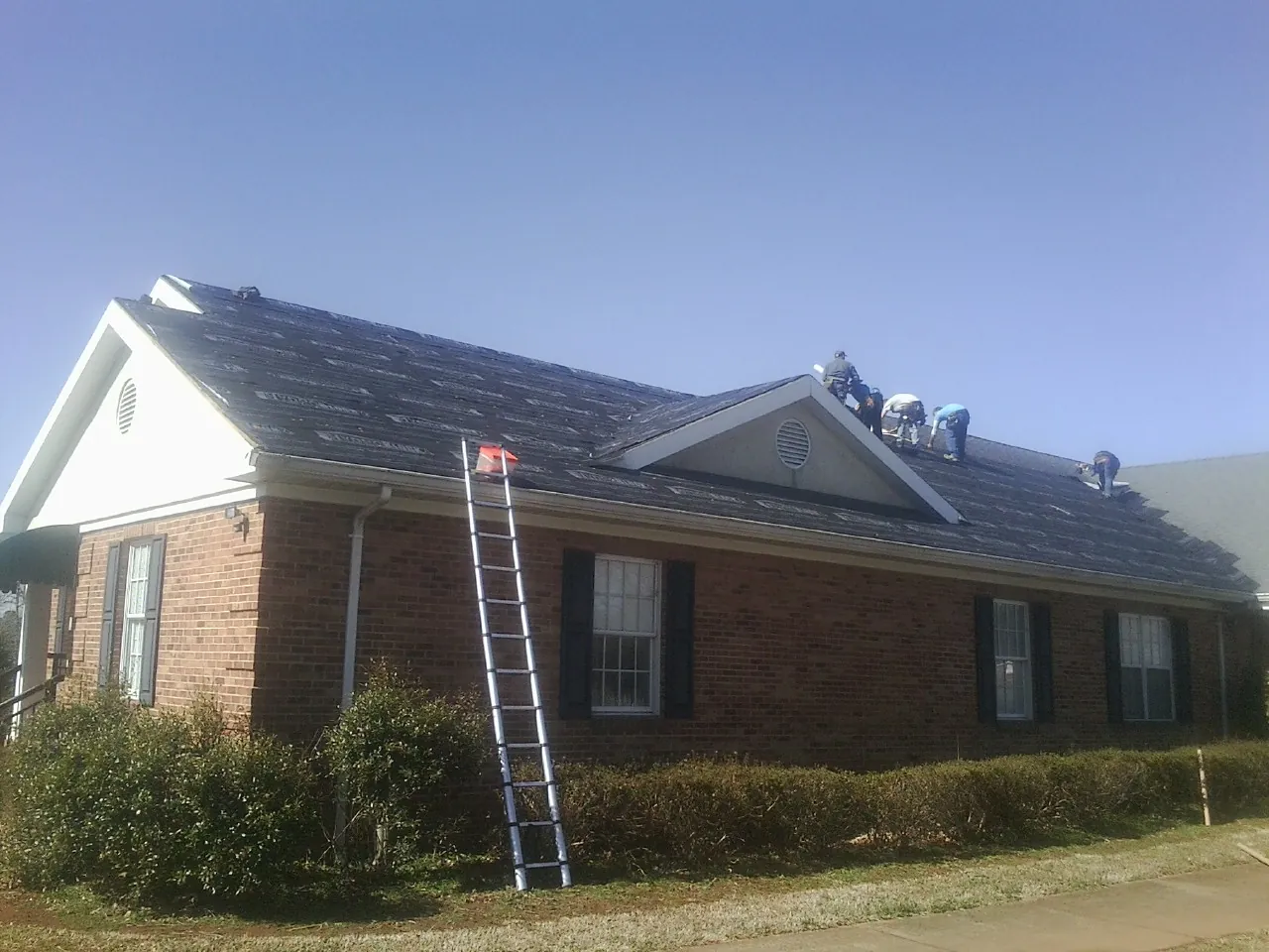Crew tearing shingles off a brick ranch roof with a ladder set — Metal Roof Repair work by our crew in Jewett