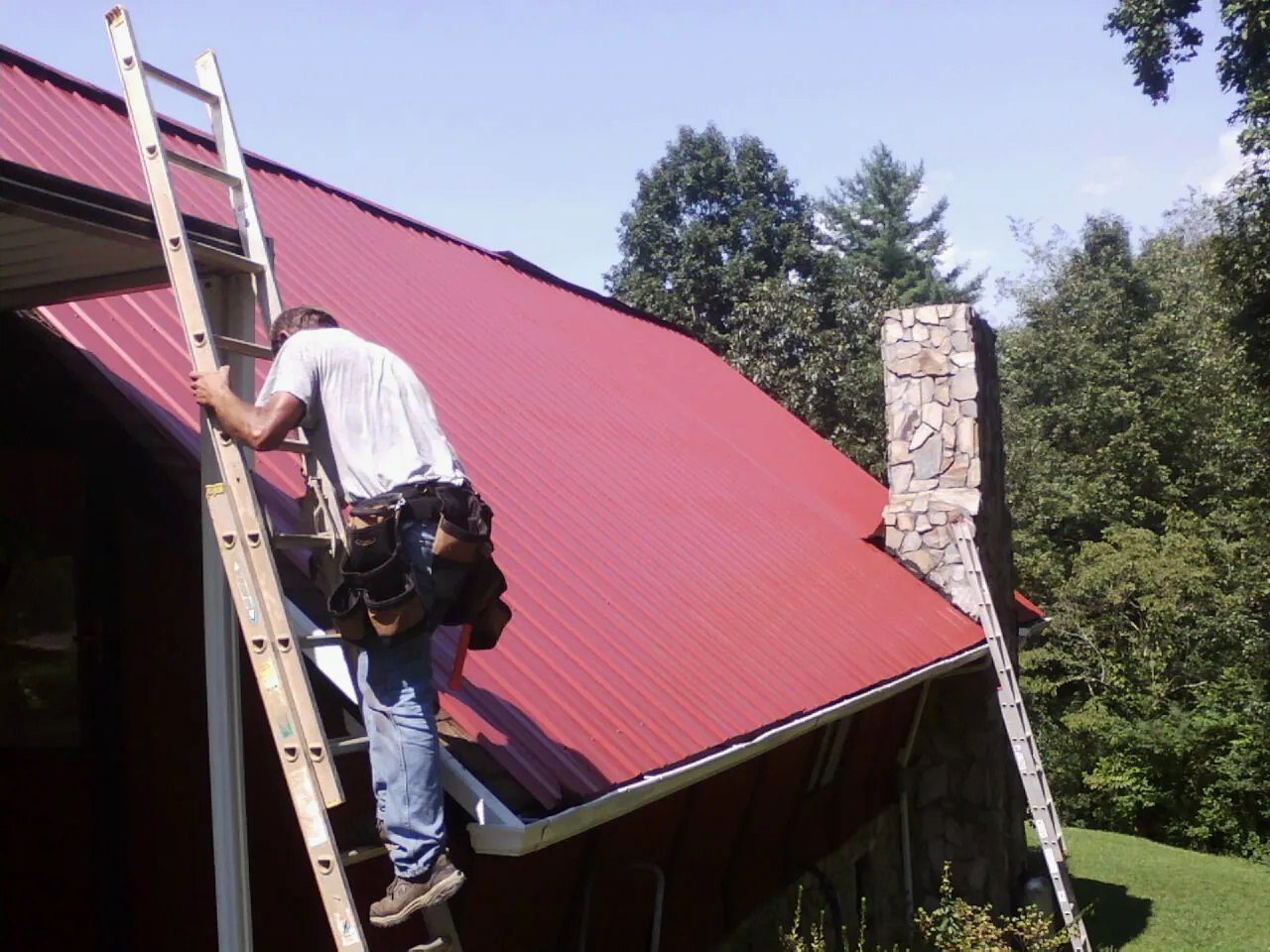 Roofer on a ladder beside a red metal roof and stone chimney — Soffit & Fascia Repair by Roofing Brothers in Jewett